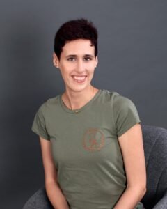 Physiotherapeutin Svenja Sabrina Hölschen. Ein Porträt einer lächelnden Frau mit kurzen, rotbraunen Haaren, die ein olivgrünes T-Shirt mit einem roten Logo trägt. Sie sitzt auf einem grauen Stuhl vor einem dunklen Hintergrund und blickt direkt in die Kamera.