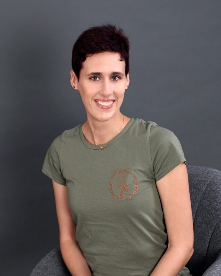 Physiotherapeutin Svenja Sabrina Hölschen. Ein Porträt einer lächelnden Frau mit kurzen, rotbraunen Haaren, die ein olivgrünes T-Shirt mit einem roten Logo trägt. Sie sitzt auf einem grauen Stuhl vor einem dunklen Hintergrund und blickt direkt in die Kamera.