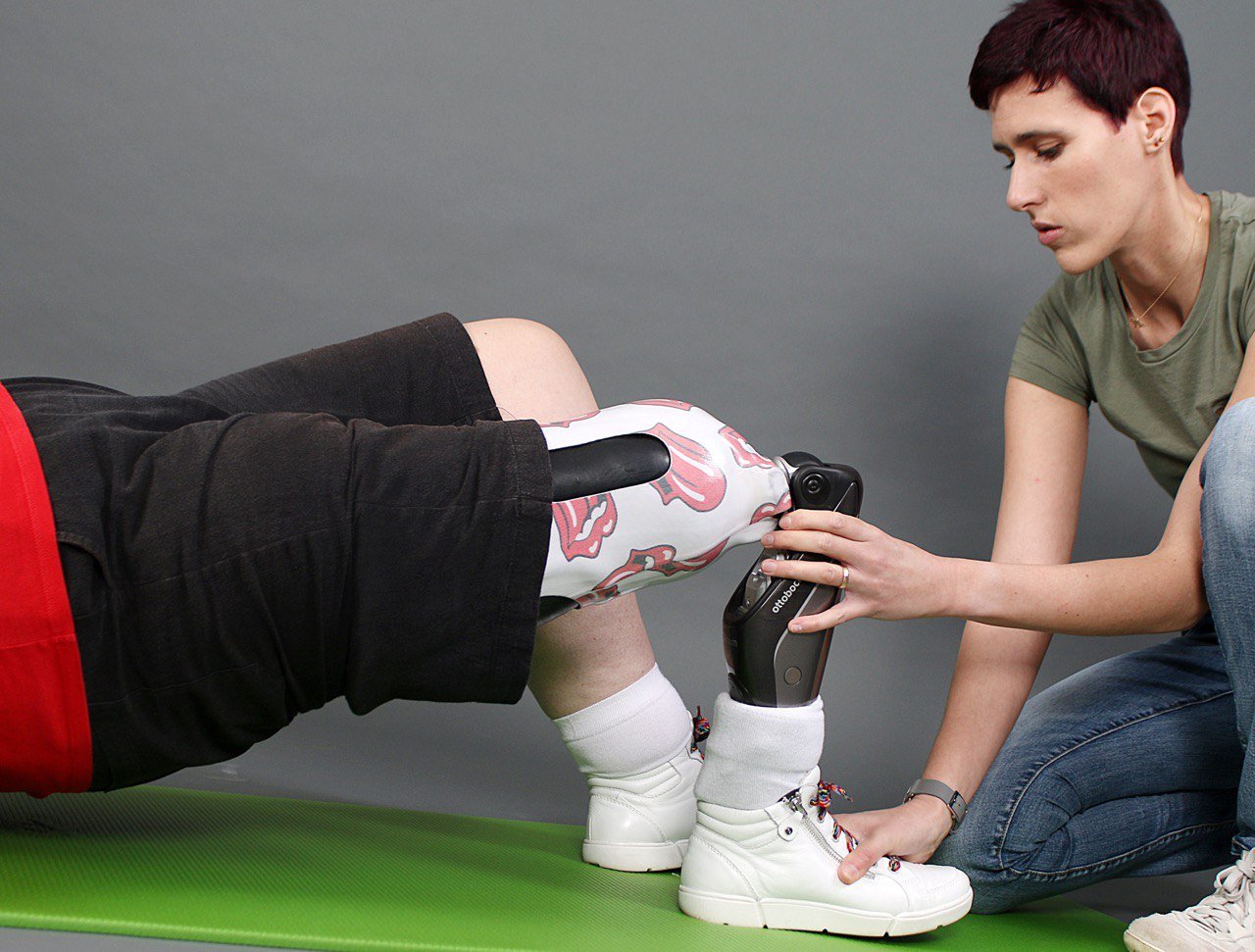 Physiotherapeutin Svenja Sabrina Hölschen. Eine Frau mit kurzen, dunklen Haaren und einem grünen T-Shirt kniet auf einem grünen Sportmatte und hilft einer Person mit einer Beinprothese, die mit einem verspielten Muster verziert ist. Die Person mit der Prothese trägt kurze schwarze Hosen und weiße Socken und Schuhe. Die Szene findet vor einem grauen Hintergrund statt.