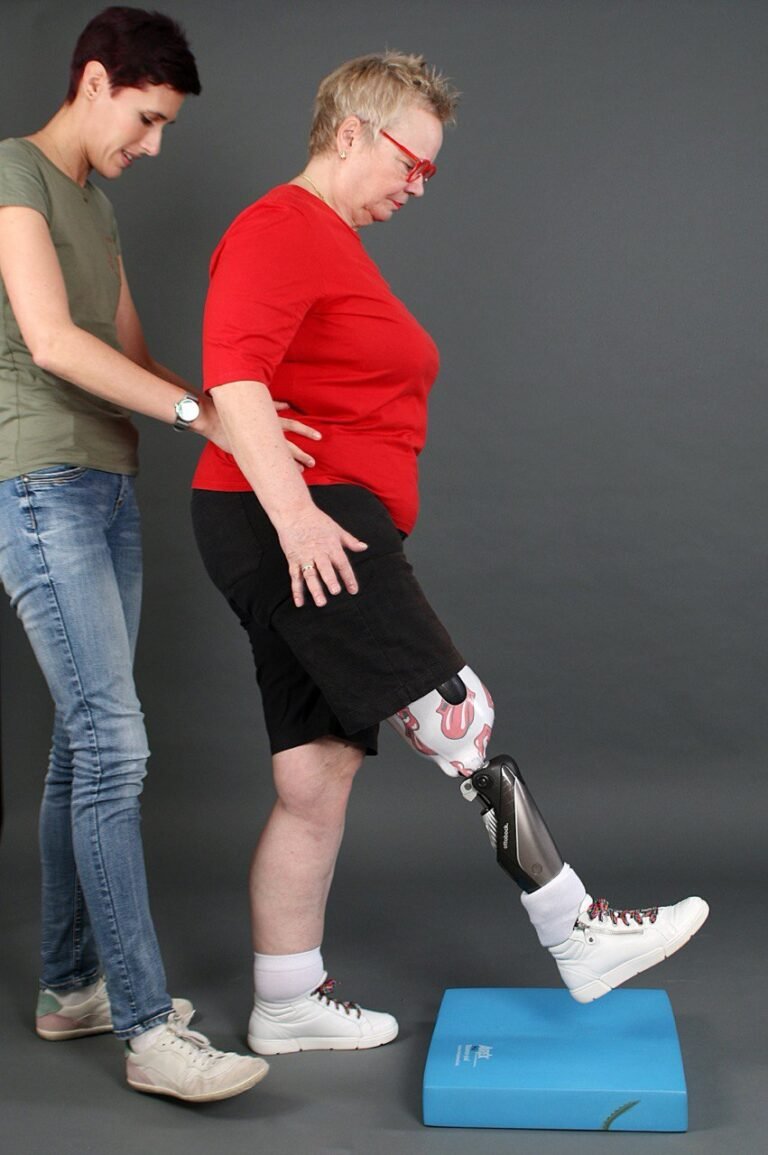Eine Pysiotherapeutin in Jeans und grünem T-Shirt unterstützt eine Patientin mit einer Beinprothese, die mit einem "Rolling Stones"-Muster verziert ist. Die Patientin, in rotem T-Shirt und schwarzen Shorts, steht mit einem Bein auf einer blauen Stufe, während die Therapeutin sie am Rücken festhält. Beide tragen weiße Sportschuhe. Der Hintergrund ist schlicht grau.